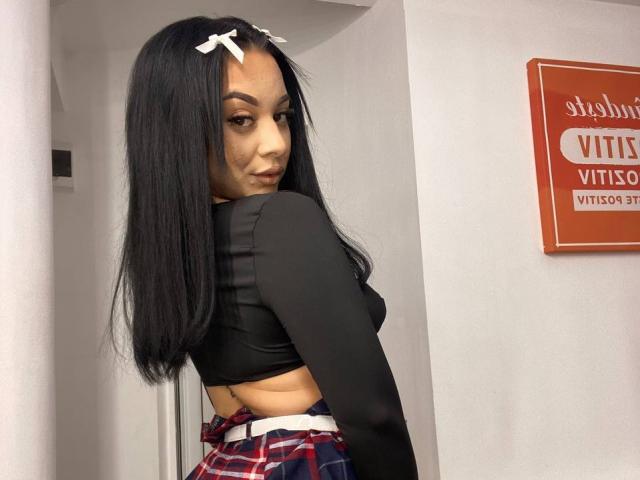 MadyLux with long black hair, white hair clips, black crop top, plaid skirt, looking over shoulder, showing her small ass and bare lower back.