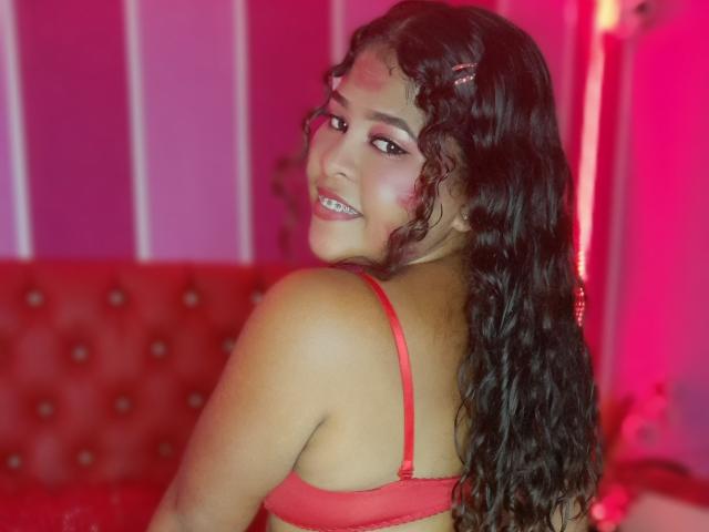 SamanthaJonesy with curly brown hair, red bra, smiling at camera, looking over shoulder, showing her tits, pink and red lighting, red tufted couch in background.