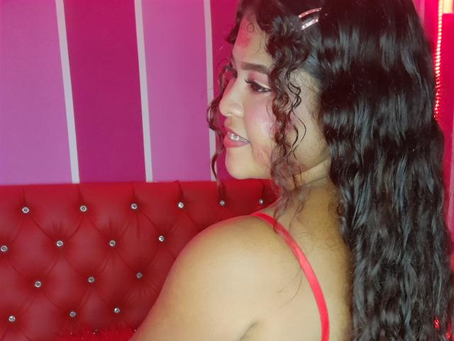 SamanthaJonesy with curly dark hair, red hairclip, red bra, smiling, showing her tits, red tufted leather couch, pink wall, red light, sexy, attractive, confident.