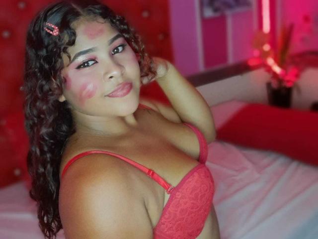 SamanthaJonesy with curly brown hair, red lace bra, pink face paint, smiling, hand in hair, bedroom, red and pink lighting, soft bed, sexy, cute, confident, flirty.