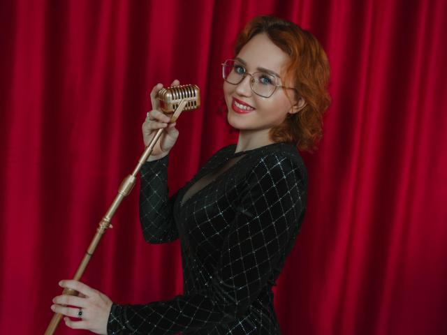 TeresaBirt with red hair, glasses, and black checkered dress, holding a golden microphone, smiling at camera, showing off her tits. Red curtain in background.