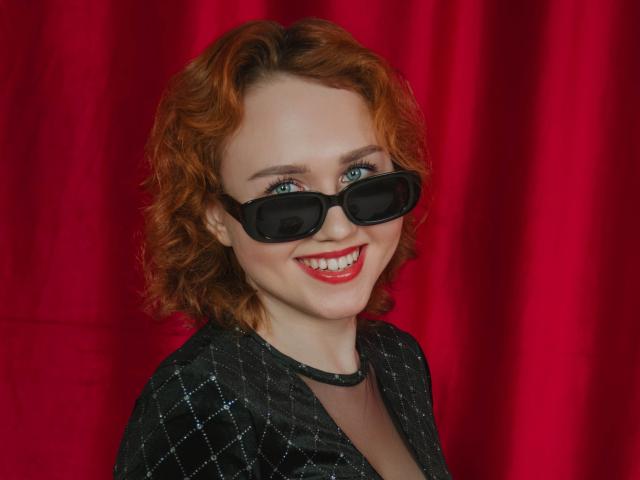 TeresaBirt has curly red hair, blue eyes, black sunglasses, and a big smile, wearing a black diamond-patterned jacket against a red curtain. Boobs are not visible.
