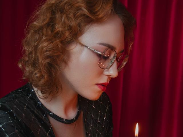 TeresaBirt with curly red hair, wearing glasses and a black, patterned top, has a serious expression, looking down at a small, glowing candle. Red curtain in background.