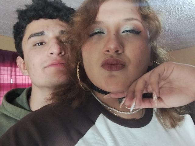 PerfectHotCouple69 with curly brown hair and lipstick, puffy eyes, and gold hoop earrings, kissing camera, wearing white and brown shirt. PerfectHotCouple69's boyfriend with curly black hair, green hoodie.