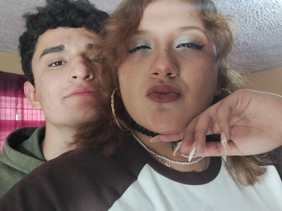 PerfectHotCouple69 with curly brown hair and lipstick, puffy eyes, and gold hoop earrings, kissing camera, wearing white and brown shirt. PerfectHotCouple69's boyfriend with curly black hair, green hoodie.