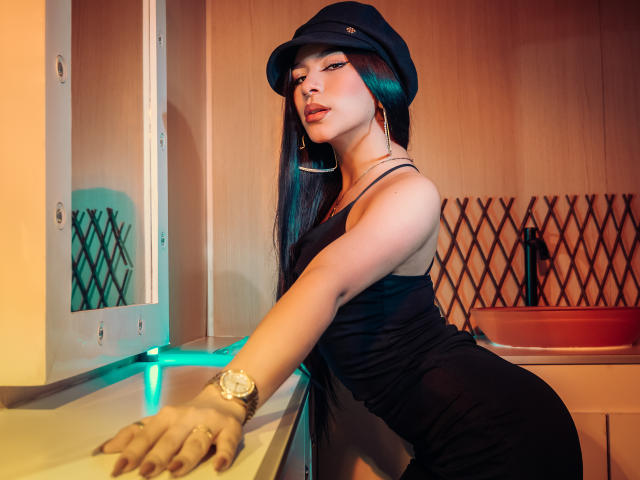 ZoeCarlter with long black hair, wearing a black cap, dress, and gold watch, leaning on a counter, looking seductively at the camera, showing her ass.