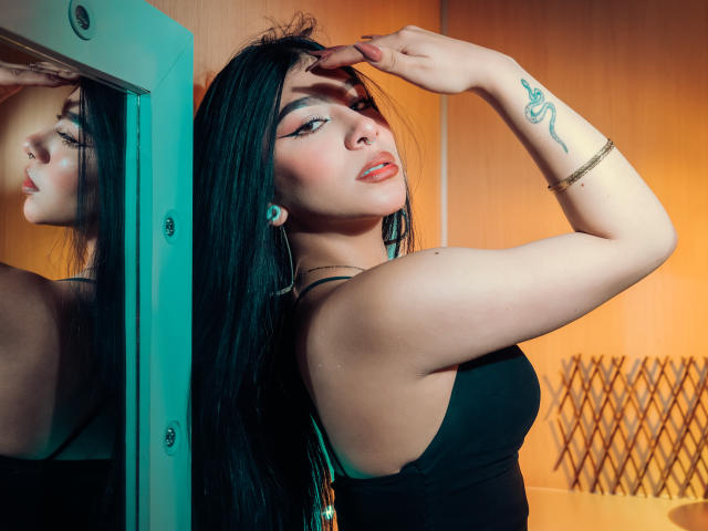 ZoeCarlter with long black hair, black tank top, and earbuds, raising her right arm, showing a snake tattoo on her wrist, looking in a mirror, with orange lighting.