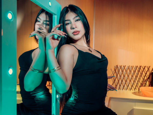 ZoeCarlter with long black hair, wearing a tight black dress, stands in front of a mirror, touching her lips, reflection showing her ass, green light on left.