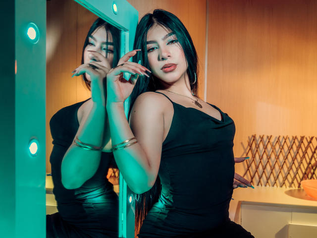 ZoeCarlter with long black hair, wearing a black dress, standing in front of a mirror, green and blue lights on, holding a pole, reflection showing her tits and face.