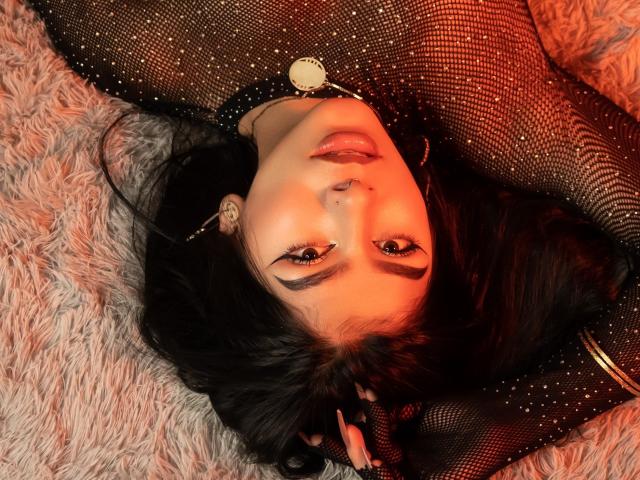 ZoeCarlter with dark hair, wearing a black fishnet dress, lying on a fluffy pink rug, red-orange light on her face, nose piercing, gold necklace, and subtle makeup.