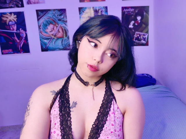 Arieyaara, con cabello negro, piel pálida y tatuajes, vestida con un top de encaje rosa, mirando hacia un lado, con lápiz labial rojo, un collar negro y un vestido de escote bajo, en una habitación iluminada con luz púrpura..
