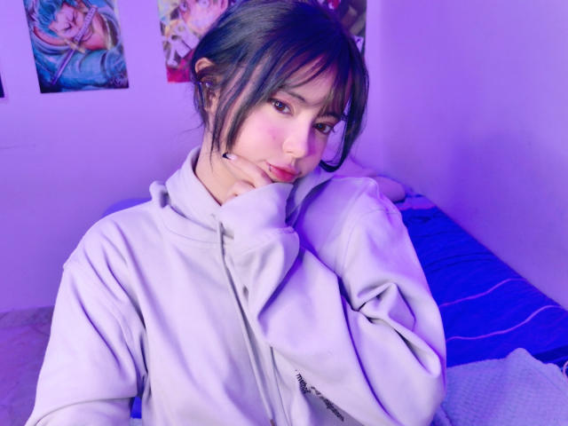 Arieyaara, com cabelo escuro, franja e bochechas rosadas, vestindo um moletom cinza claro, tocando o queixo, em um quarto iluminado em tons de roxo, com pôsteres de anime ao fundo..
