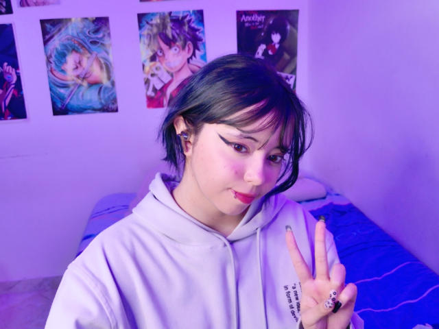 Arieyaara with black hair, white hoodie, and pink eyeliner, showing a peace sign, has a ring on her finger and purple-tinted room with anime posters.