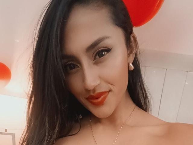 Helen Madisson tiene el pelo oscuro, lápiz labial rojo y lleva un collar con pequeñas perlas. Está sonriendo y tiene un piercing en la nariz y pendientes de diamantes. En el fondo, hay globos rojos..