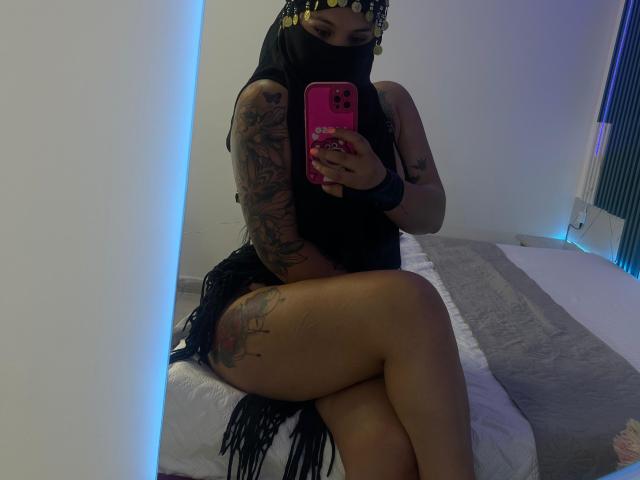 MalakCote with tattoos, wearing black veil and fringed dress, holding pink phone, sitting on bed, showing bare ass and legs, looking at mirror, dim blue-lit room.
