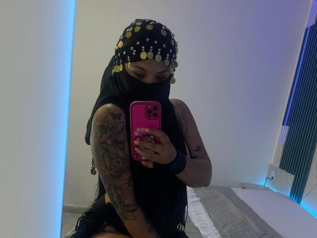 MalakCote with tattoos, wearing a black mask, black headscarf with gold coins, black sleeveless top, holding pink phone, taking mirror selfie, visible arm tattoos, sitting on bed.
