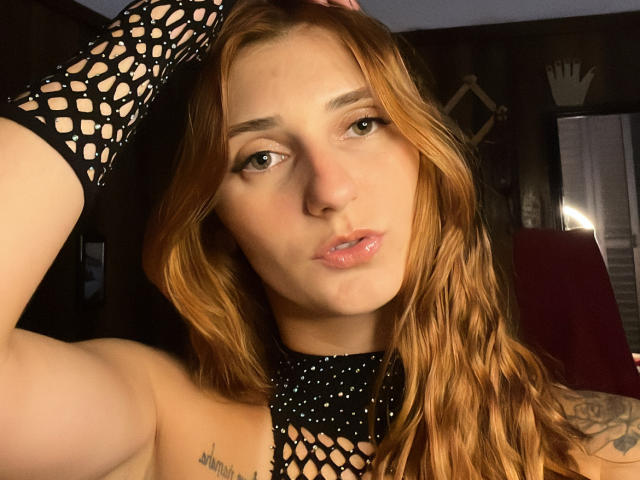 Melissadry with wavy red hair, wearing a black fishnet sleeve, pouty lips, and a tattoo on her left shoulder, showing off her tits and a confident, sexy look.