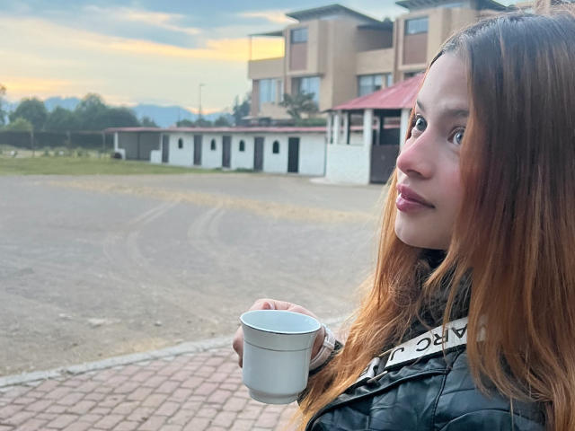 Shantal, con lunghi capelli castani, tiene in mano una tazza bianca, indossa un giubbotto nero imbottito, è in piedi all'aperto e guarda di lato con un sorriso appena accennato, con il tramonto sullo sfondo..