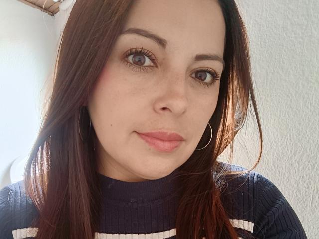 Le visage de Seher, avec ses cheveux bruns, ses yeux bleus et ses lèvres roses, porte un pull marine foncé avec des rayures blanches. Elle regarde l'appareil photo avec un léger sourire..