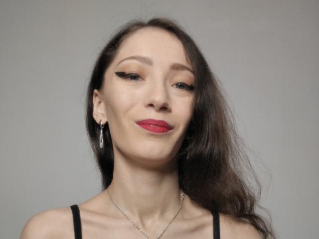 Silerta has long brown hair, red lipstick, and black eyeliner, wearing a black top, silver earrings, and a necklace, with a slight smile, looking confident.