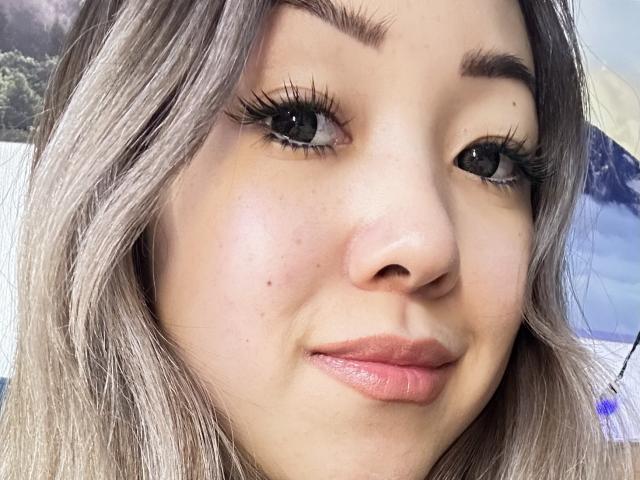 RenyLime's face with light brown, wavy hair, full lips, and dark, long eyelashes. She has a small mole on her right cheek and is smiling slightly.