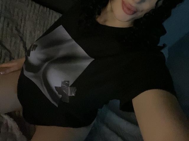 Olieri's black shirt with gray and white cross, showing a slinky, curvy, pale-skinned butt, and a slightly visible, round pussy, with dark curly hair and pouty lips.