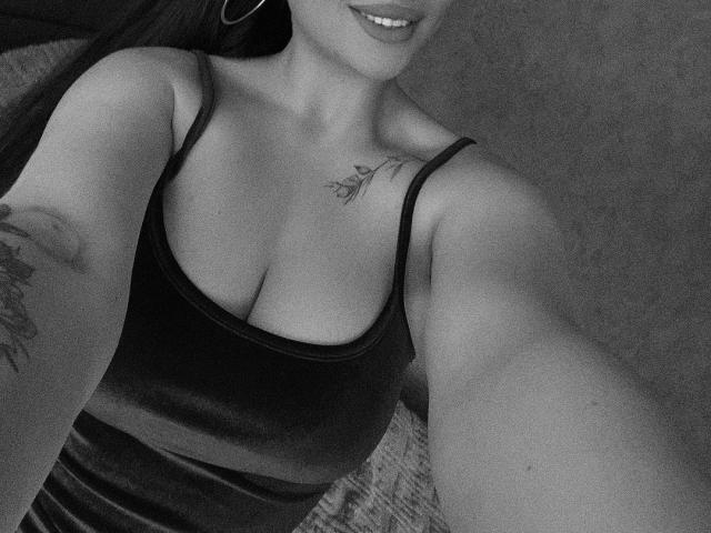 Olieris schwarzes Tanktop enthüllt ihre Brüste, mit einem kleinen Tattoo an ihrer rechten Schulter und einem weiteren an ihrem linken Arm. Sie lächelt und trägt Ohrringe im Stecker-Stil. Schwarz-weißes Selfie..