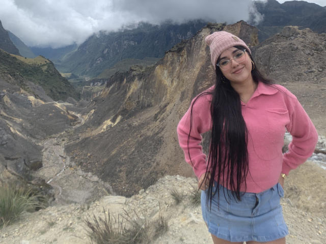 VictoriaSaens with long black hair, pink beanie, glasses, pink jacket, and blue skirt, standing in a rocky mountain landscape, smiling, hands on hips, showing her ass.