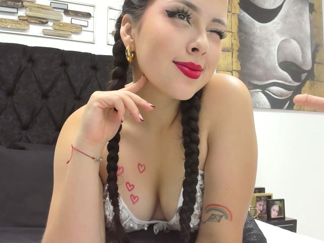AlisonTatcher with braided hair, winking, red lipstick, gold earrings, pink hearts tattoo on chest, rainbow tattoo on arm, winking, pink nails, black bed, white wall, black headboard.