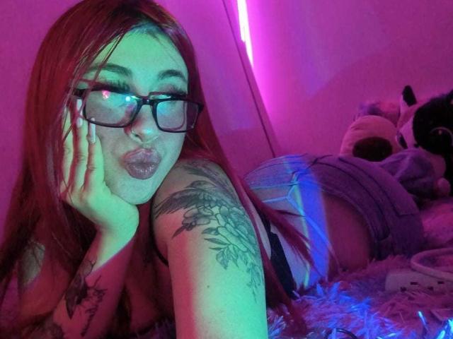SammyViolet with long red hair, glasses, pouty lips, tattooed arm, lying on bed, wearing denim shorts, pink and blue neon light, hand on cheek, sexy, cute, fluffy blanket.