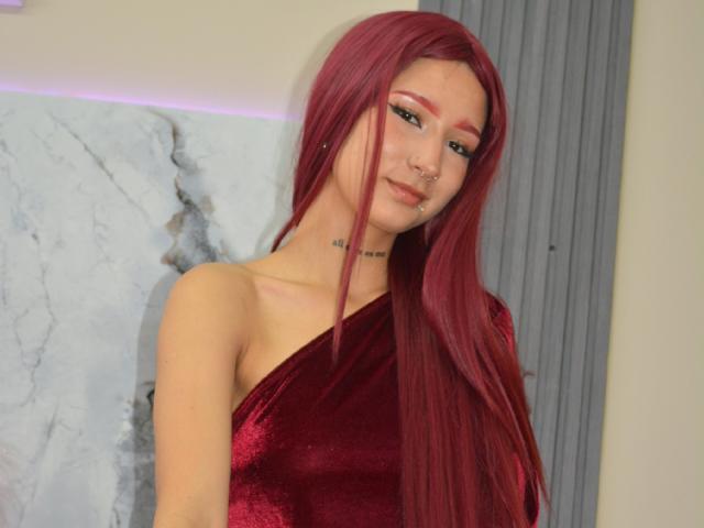 CathaLennon with long red hair, wearing a red velvet one-shoulder dress, smiling with a nose ring and eyeliner, looking cute and sexy.