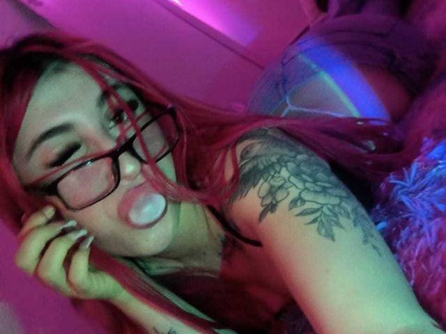 SammyViolet with red hair, glasses, and a blow job face, showing her tattooed arm and puffy ass in pink and blue lighting. Her tits are partially visible.