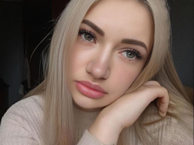 MikkiShows has pale skin, blonde hair, and green eyes. She's looking at the camera with a slight smile, head resting on her hand, wearing a pinkish sweater.