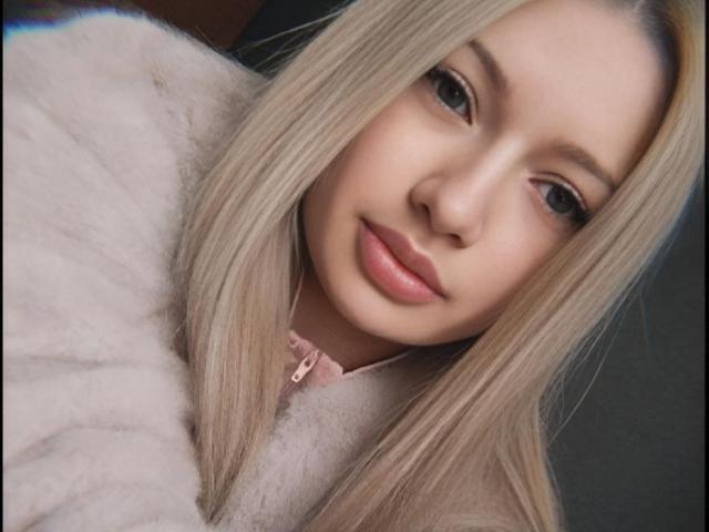MikkiShows has blonde hair, pink lips, and soft-looking skin. She's wearing a fluffy pink top, looking straight at the camera with a slight, cute smile.