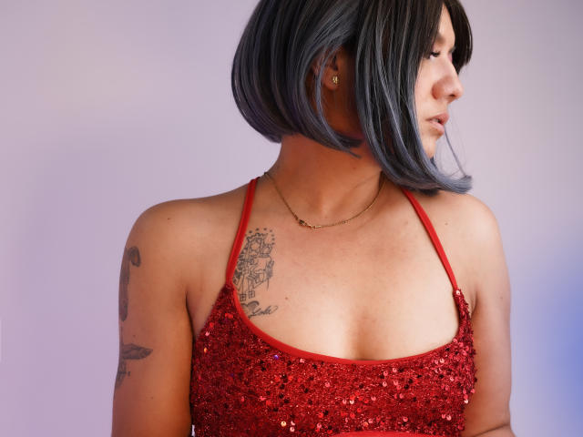 ColetteM has a shiny red sequined top, black bob with blue highlights, and tattoos on her left shoulder and right chest. She's wearing a gold necklace.