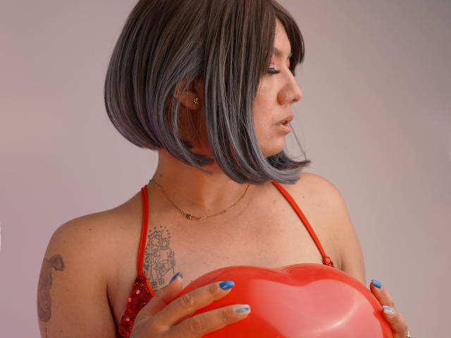 ColetteM mit einem Bob-Haarschnitt, blauen Nägeln, einem roten Top mit Pailletten, die einen roten Herzballon hält, ein Tattoo an der rechten Schulter, eine goldene Halskette, Profilansicht, Blick nach rechts..