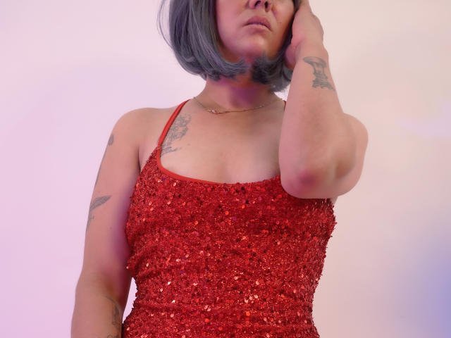ColetteM trägt ein rotes, paillettenbesetztes Kleid, hat eine Hand am Hals, einen grauen Bob, Tattoos am Arm, eine goldene Halskette um den Hals, dezentes Make-up, eine mittelgroße Brust und leicht geschwollene Lippen..