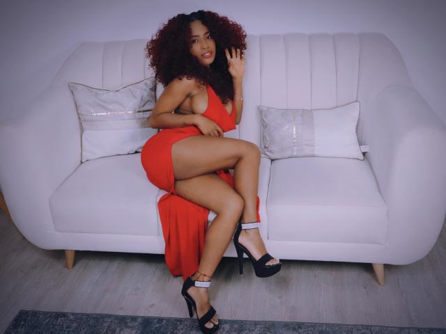 Ameli Bernard, con i capelli rossi ricci, indossa un vestito rosso, tacchi alti neri, è seduta su un divano bianco, mostra il sedere e le gambe, sorride e saluta..