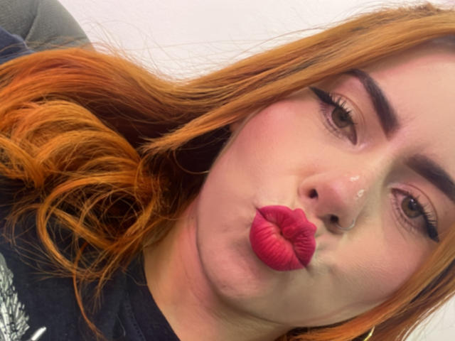 DulceCharlotteHot with orange hair, thick eyebrows, and pink lipstick, pucker her lips with a white fluid on her nose, wearing a black top, in a selfie.