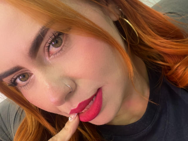 DulceCharlotteHot with orange hair, brown eyes, red lipstick, nose ring, and gold hoop earrings, touching her lip with a finger, wearing a black shirt.