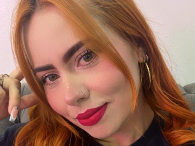 DulceCharlotteHot has orange hair, brown eyes, thick eyebrows, red lipstick, gold hoop earrings, and a nose ring. She's smiling with her hand behind her head.