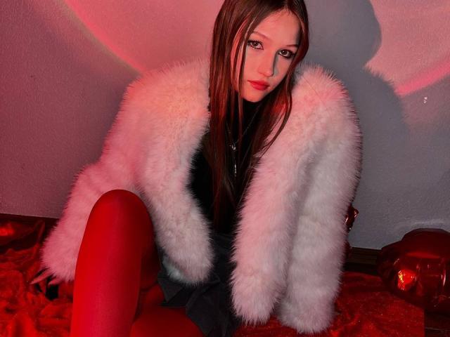 Bubochka with long brown hair, red lips, wearing a fluffy white coat, black turtleneck, and red tights, sitting on red fabric, looking at camera, necklace with cross.