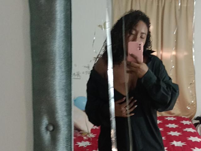TaniaCooper69, oben ohne, mit langen braunen Haaren, zeigt ihren Hintern und einen kleinen schwarzen String, blickt über ihre Schulter und steht in einem Raum mit einer Holzbalkendecke..
