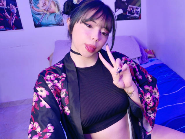 Arieyaara with black pigtails, tongue out, wearing a floral kimono, black choker, and ribbed black crop top, flashing a peace sign with a ring, in a purple-lit room.