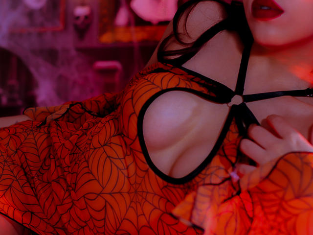 HottSirenn's red, spider-web patterned dress reveals her big, round tits with a black strap, her hand gripping the fabric, and her mouth slightly open. Red lighting.