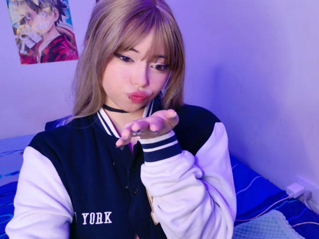 Arieyaara with blonde hair, black choker, and a black-and-white varsity jacket, puckering her lips and pointing at the camera, showing her ringed finger. Blue-purple light.