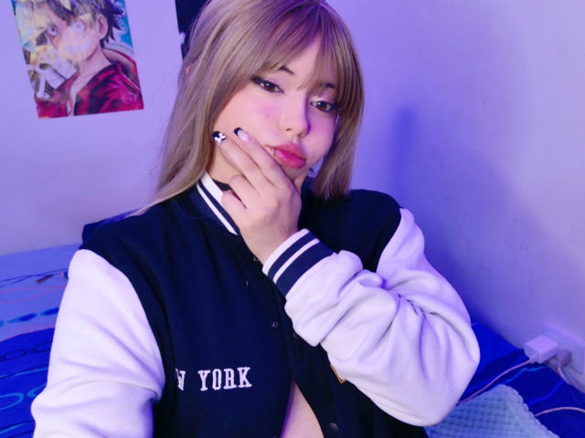 Arieyaara with blonde hair, wearing a black and white varsity jacket, touching her cheek with a hand, showing pink blush on her face, sitting in a blue-lit room.