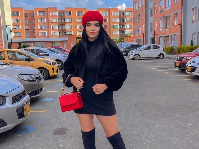 NovaDixon, con lunghi capelli neri, un berretto rosso, una giacca di pelliccia nera, un vestito nero, una borsa rossa e stivali neri, è in piedi in un parcheggio con auto gialle e argentate..
