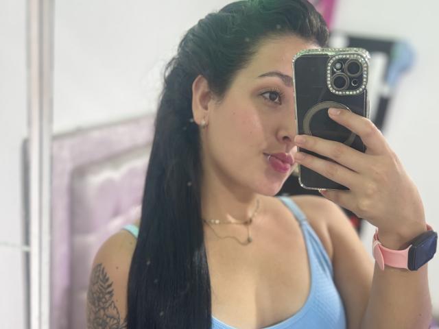 SarahGood with long black hair, pouty pink lips, blue tank top, pink watch, tattoo on left shoulder, taking a mirror selfie, holding a phone with a black case.