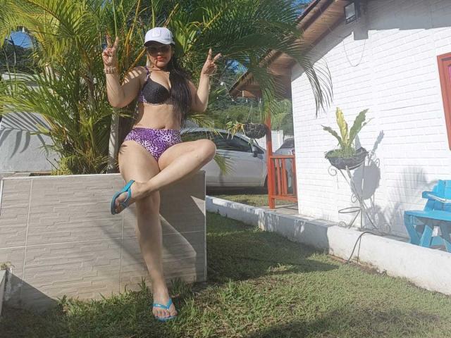 KinkyVane with long dark hair, wearing a purple and black bikini, purple high-waisted bottoms, blue flip-flops, white cap, making double peace signs, standing in front of a white house.